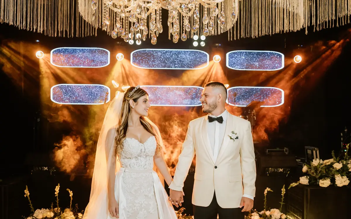 Novios tomados de la mano en una pista blanca, bajo una lámpara de cristal y cortinas de flecos